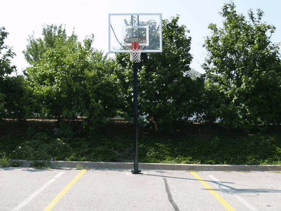 A basketball hoop is mounted on a black pole in the middle of a parking lot.
