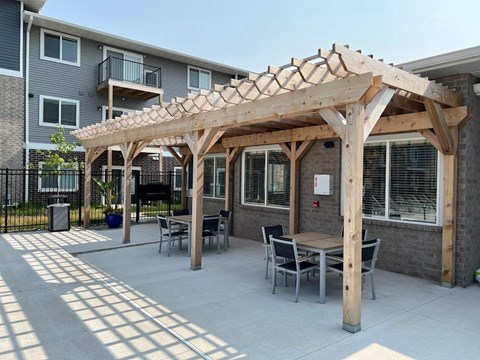 A wooden pergola with a table and chairs is in front of a building.