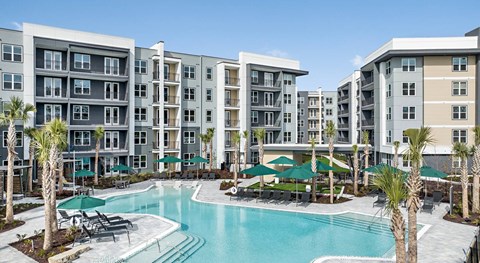 A large swimming pool surrounded by lounge chairs and umbrellas in front of apartment buildings.