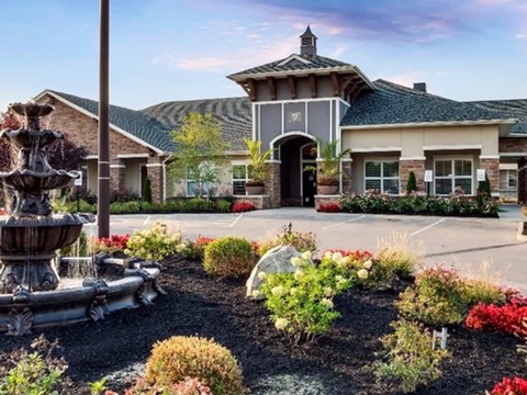 A fountain is in the middle of a garden with a house in the background.