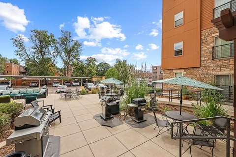 Peregrine Place - Patio with a grill and chairs is surrounded by a building and trees.