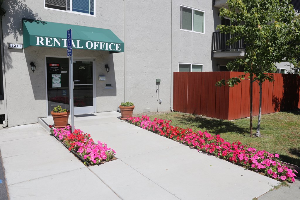 The front entrance of the Aegena apts, the entrance to the office lined with pink flowers and a green banner Prodesse Property Group 1211 Garbo Way  San Jose, CA 95117