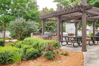 A wooden pergola with a sign on it is surrounded by greenery and a picnic table.