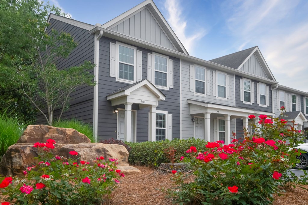 A house with grey siding and white trim is surrounded by red flowers and greenery at Ansley at Town Center Apartments, Evans, Georgia
