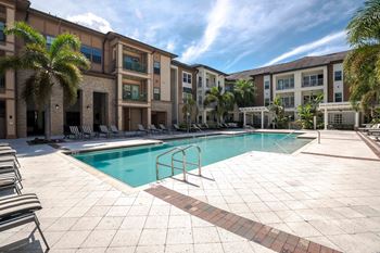 A swimming pool surrounded by a tiled patio and lounge chairs in front of a building with palm trees at Park Place Oviedo Apartments, Oviedo