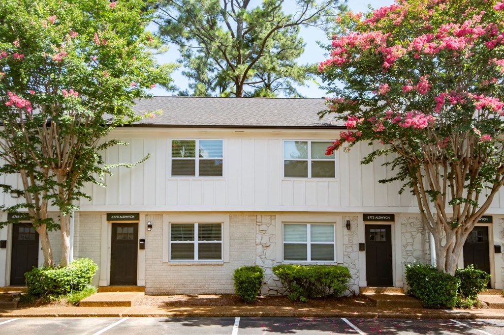 A white building with black doors and windows is surrounded by greenery and trees.