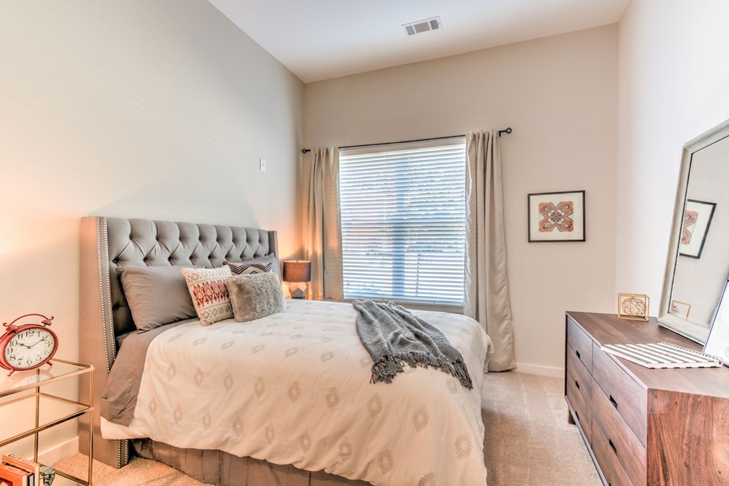 A bedroom with a large bed, a nightstand with a clock, a dresser with a mirror, and a window with curtains at Highland Row Apartments, Tennessee