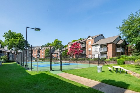 A tennis court is surrounded by a green lawn and apartment buildings.
