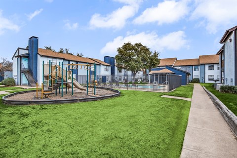 A playground with a slide and swings is surrounded by a fence and a grassy area.