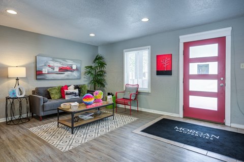A living room with a grey couch, a coffee table, a red door, and a painting on the wall.