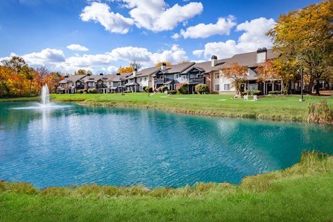 A serene lake with a fountain in the middle surrounded by a grassy area and buildings in the background.