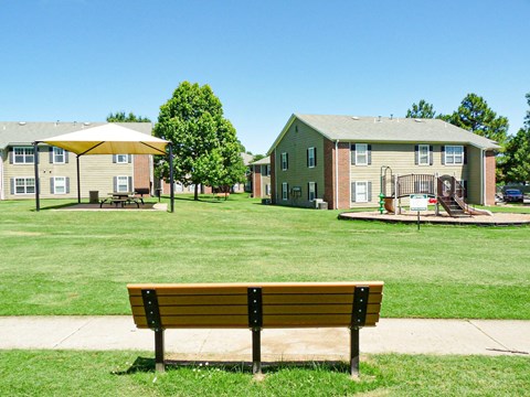 A park bench sits in the foreground of a grassy area with apartment buildings in the background.