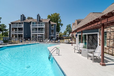 A large swimming pool is surrounded by lounge chairs and a building with a balcony overlooks the pool.