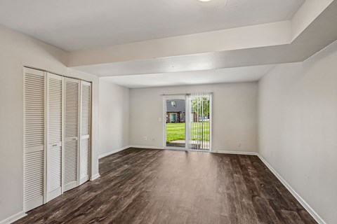 A room with wooden floors and white walls, with a view of a green lawn and a building through a glass door.
