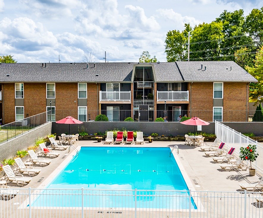 A large swimming pool in front of a brick building with red umbrellas and lounge chairs.