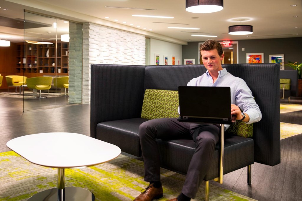 A man in a white shirt and black pants is sitting on a black couch with a laptop on his lap.