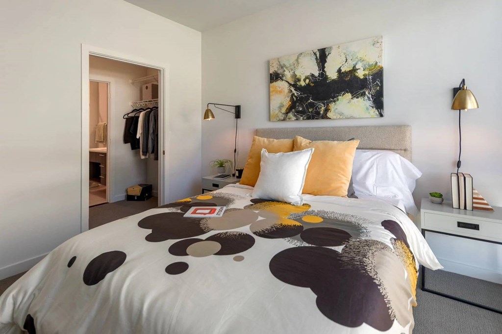 A bed with a white comforter and a brown and black pattern is in the foreground of a room with a painting on the wall and a clothing rack in the background.