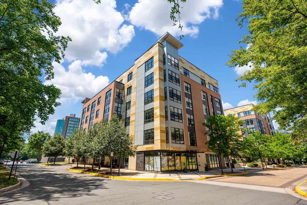 A modern multi-story building with a mix of brown and beige colors is surrounded by trees and a clear sky.