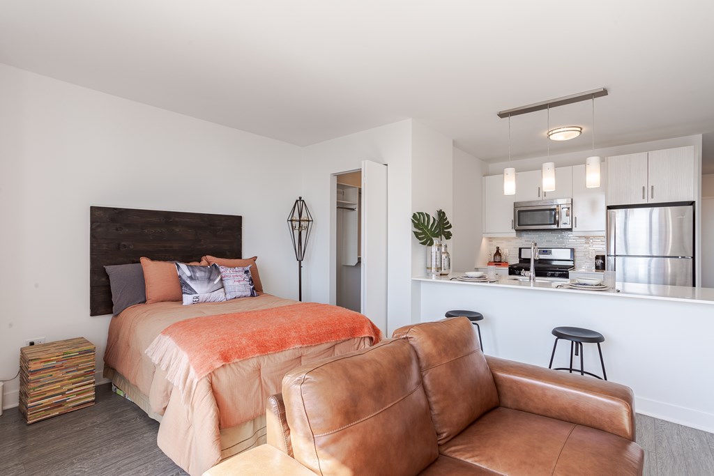 A bedroom with a bed, a couch, and a kitchen area in the background.