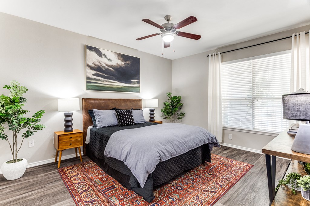 Dog-Friendly Apartments in North Austin, TX - Sycamore Springs - Furnished Bedroom with a Ceiling Fan, a Large Window, and Faux Plank Flooring