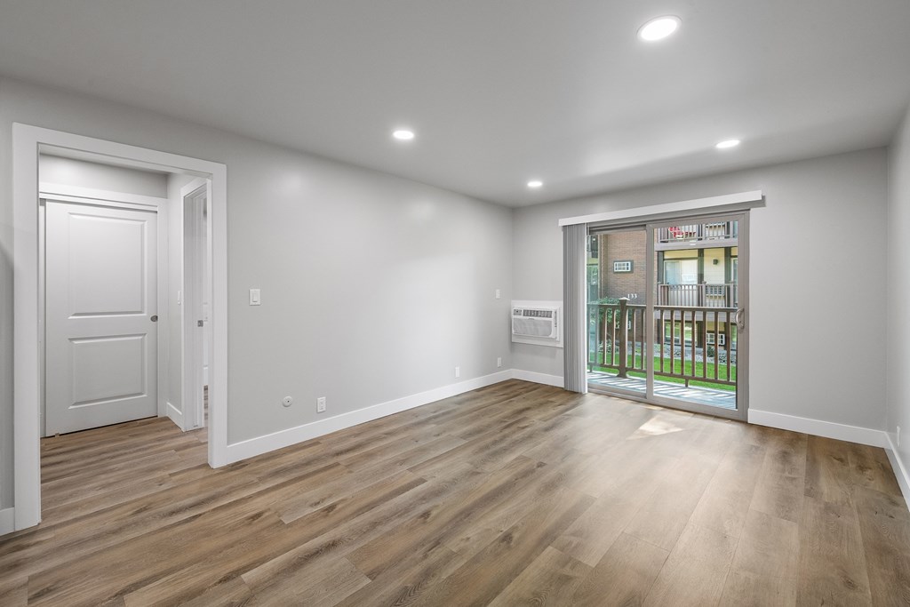 A room with wooden floors and white walls, with a door on the left and a balcony on the right.