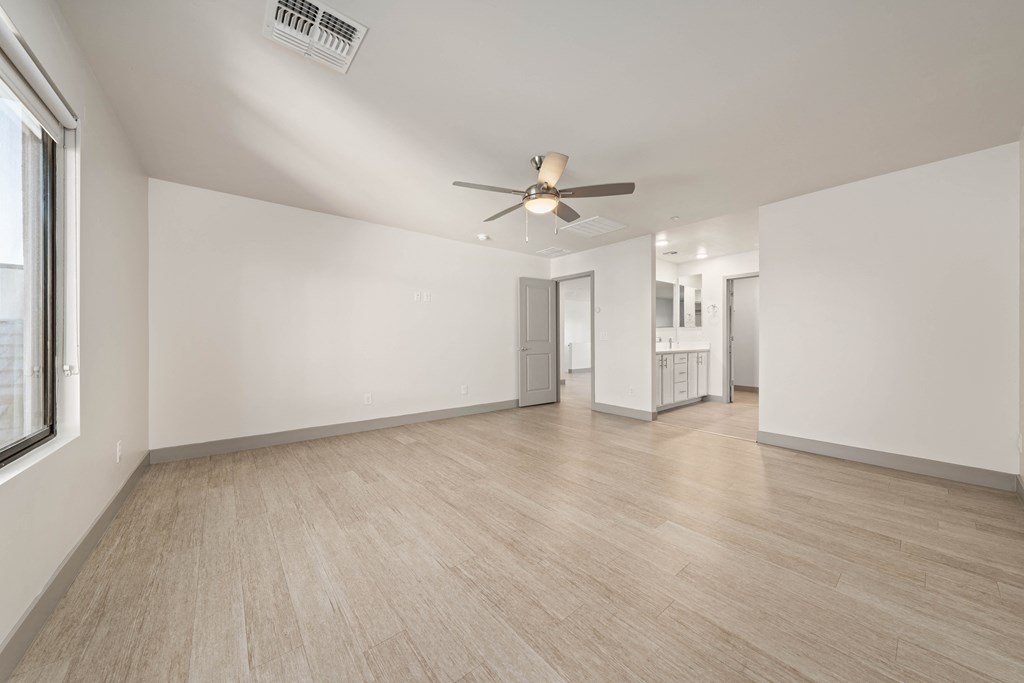 A spacious room with a ceiling fan and light, hardwood flooring, and a window on the left side.
