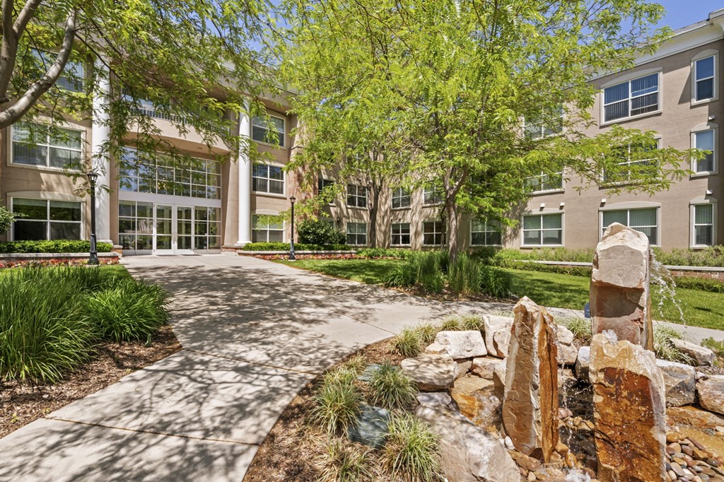 a pathway leading to an apartment building with trees and a rock sculpture in the foreground
