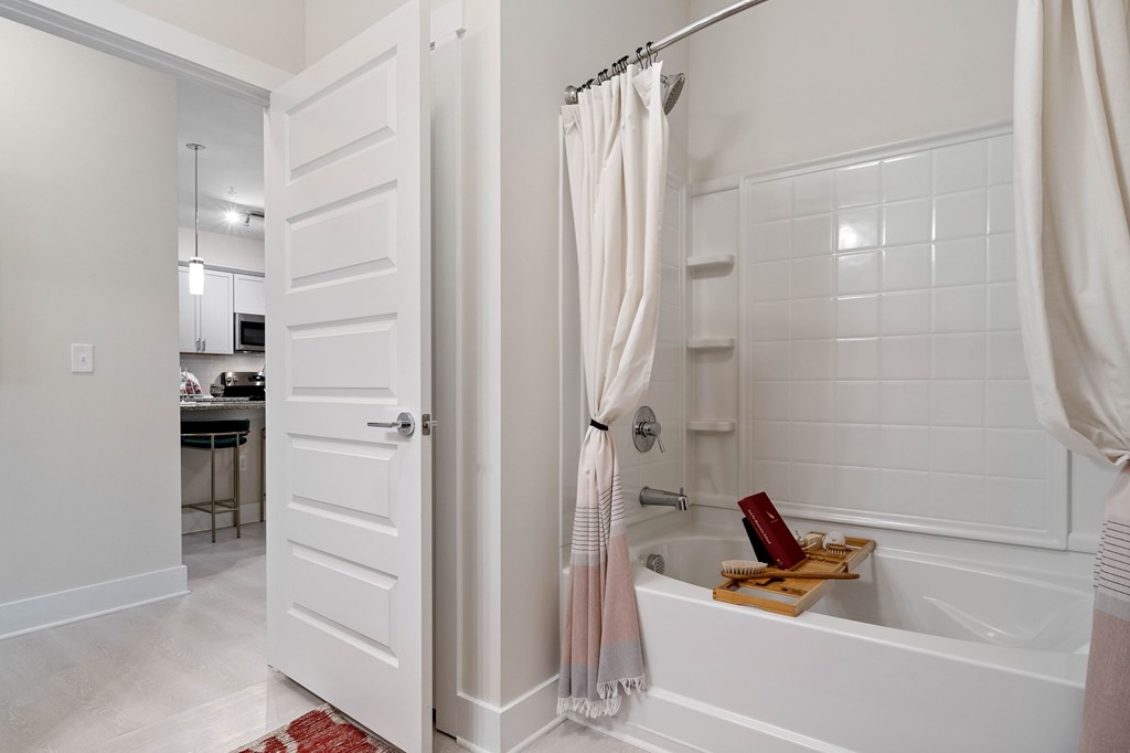 A white bathroom with a tub, shower curtain, and a red rug.
