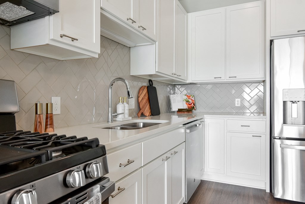 a kitchen with white cabinets and a stove and sink Hopkins, MN Luxury Apartments Near Methodist Hospital - The Hallon - Modern Kitchen with White Cabinetry, White Countertops, White Tile Backsplash, a Large Cooking Island, Hanging Lights, Stainless Steel Appliances, and Wood-Finish Plank Flooring