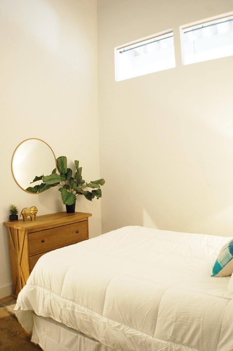 A bedroom with a white bed and a wooden dresser with a round mirror and a plant on top.