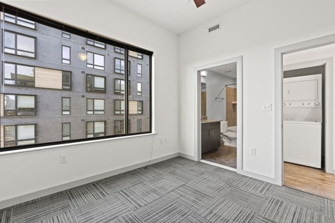 A room with a grey carpet and a large window overlooking a building.