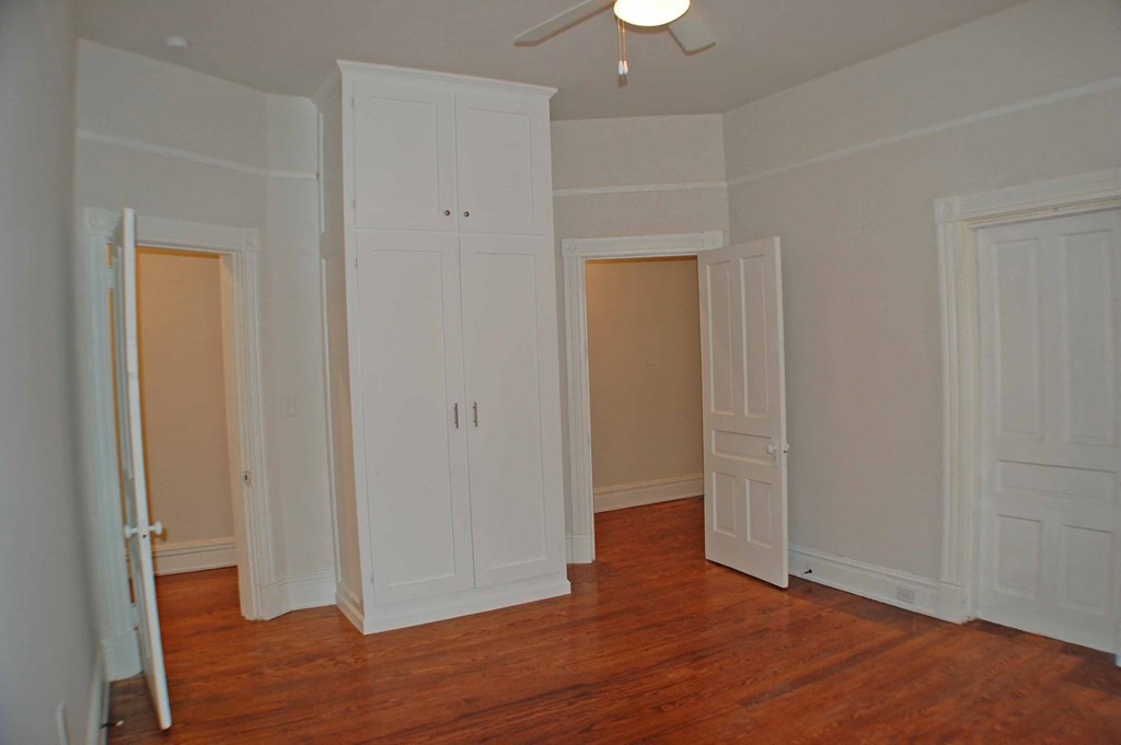 A room with white walls and wooden floors has a ceiling fan and three white doors.