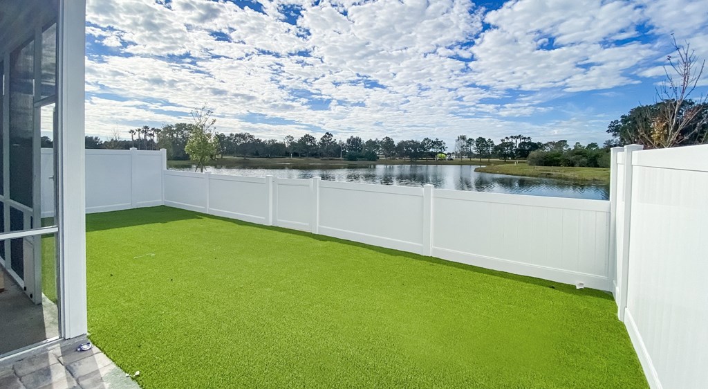 A white fence surrounds a green lawn with a lake and trees in the distance.