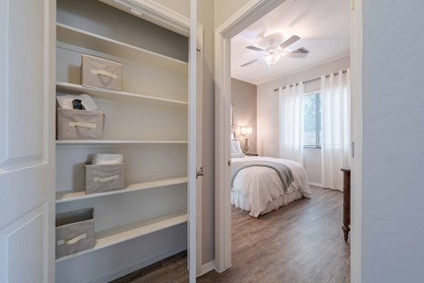 A white closet with shelves and drawers is open to a bedroom with a bed and a ceiling fan.