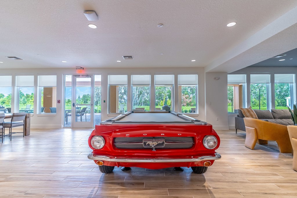 A red vintage car is parked in a spacious room with wooden floors and large windows.