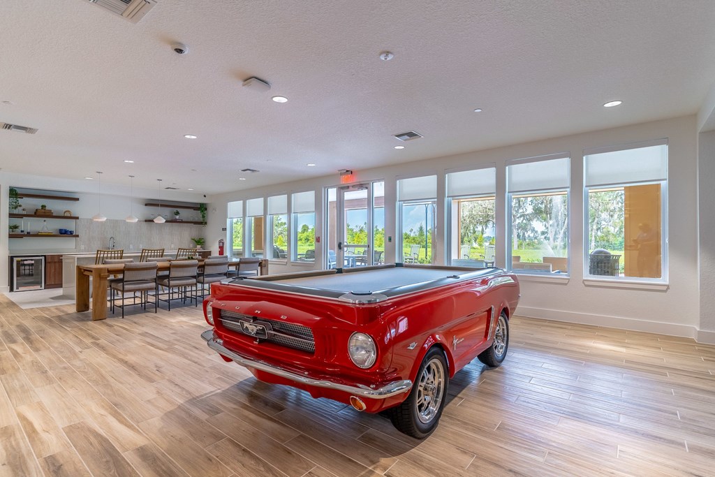 A red vintage car is parked in a spacious room with wood flooring and large windows.