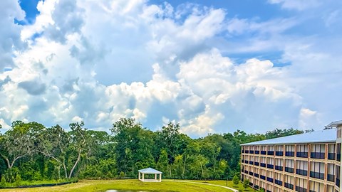 A large building is situated on a grassy area with a gazebo and trees in the background.
