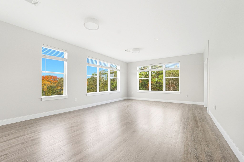 A spacious room with wooden flooring and three windows showing autumn trees outside.