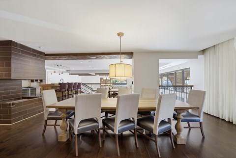 Long clubroom dining table seating ten under a drum pendant, with a tile fireplace on one side and a bar beyond.