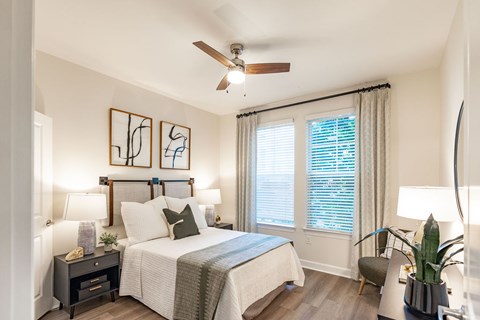 Bedroom with a ceiling fan, two windows with blinds and curtains, nightstand lamps, and wood-look plank flooring.