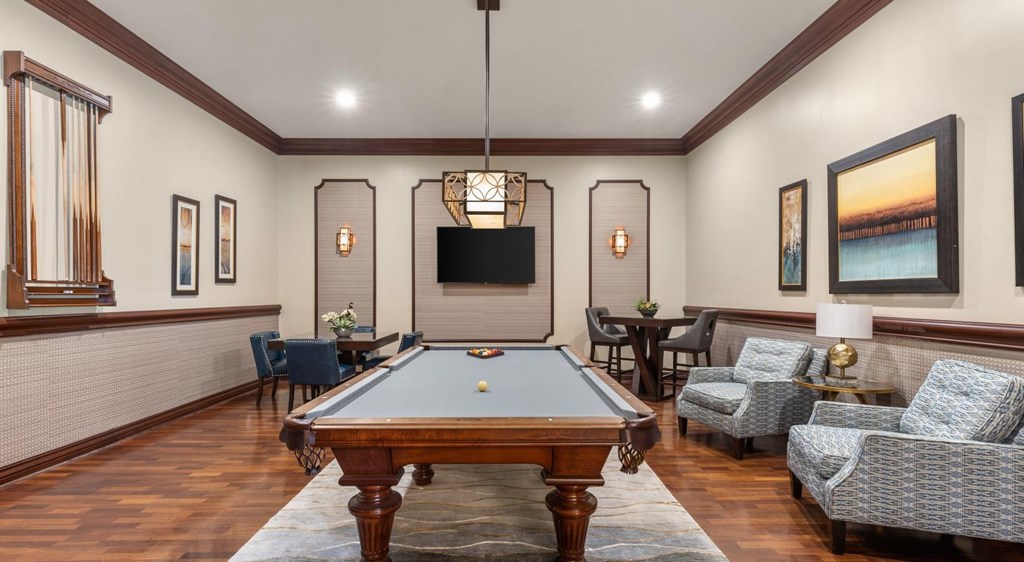 A pool table is surrounded by chairs in a room with a television and pictures on the wall.