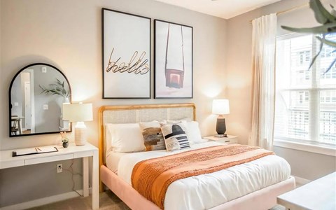 Bedroom with a cane headboard, framed prints, matching nightstand lamps, and a white desk beneath an arched mirror.
