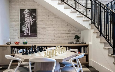 Game area beneath the clubhouse staircase with a chess table, white chairs, framed artwork, and a low bookshelf.