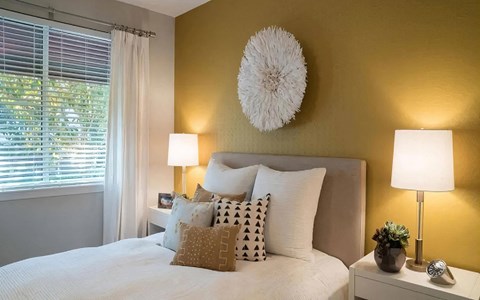 Bedroom with an upholstered headboard against a gold accent wall, matching nightstand lamps, and a curtained window.