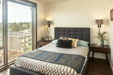 Bedroom with corner floor-to-ceiling windows, a tufted upholstered headboard, wall sconces, and walnut nightstands.