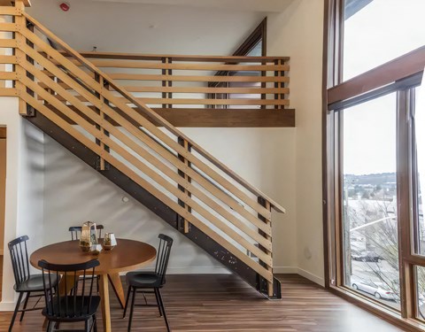 Loft interior with a steel and wood staircase, a round dining table with black chairs, and tall wood-framed windows.