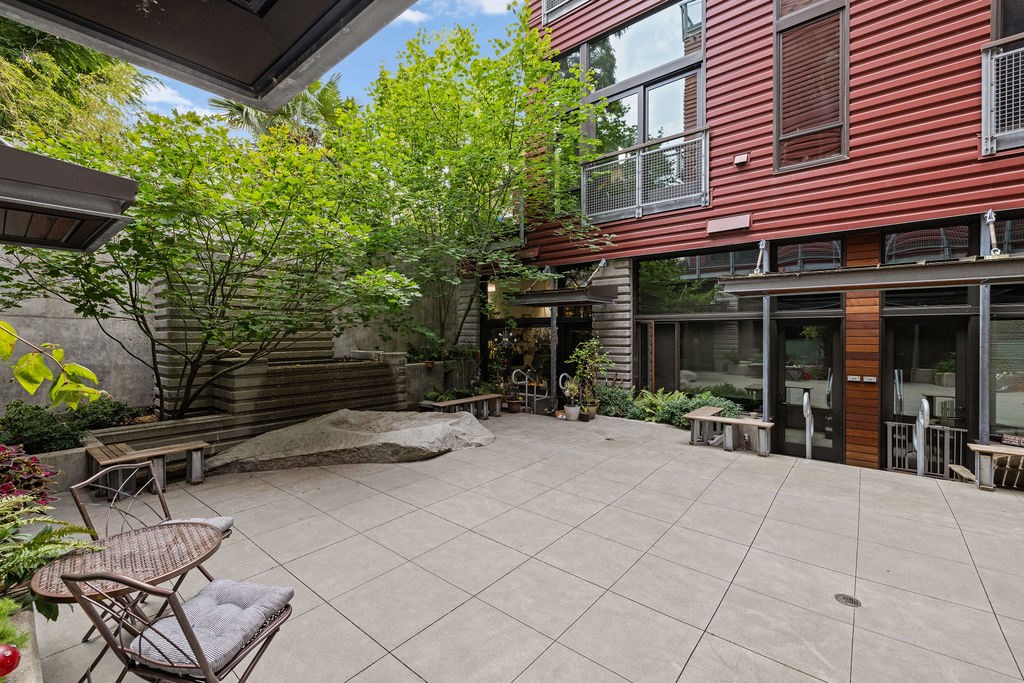 A patio with a table and chairs is surrounded by a red building and trees.