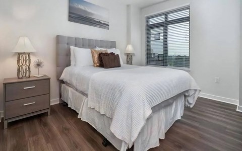 Bedroom with a gray tufted headboard, framed seascape, a nightstand lamp, and a blinded window beside the bed.
