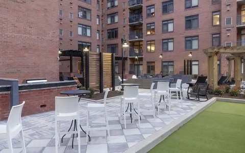 Courtyard with high-top tables and white bar chairs on checkerboard paving, with a turf strip and pergolas behind.