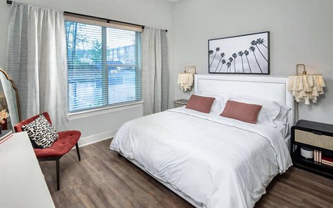Apartment bedroom sized for a king bed, with a wide window, upholstered headboard, accent chair, and plank flooring.